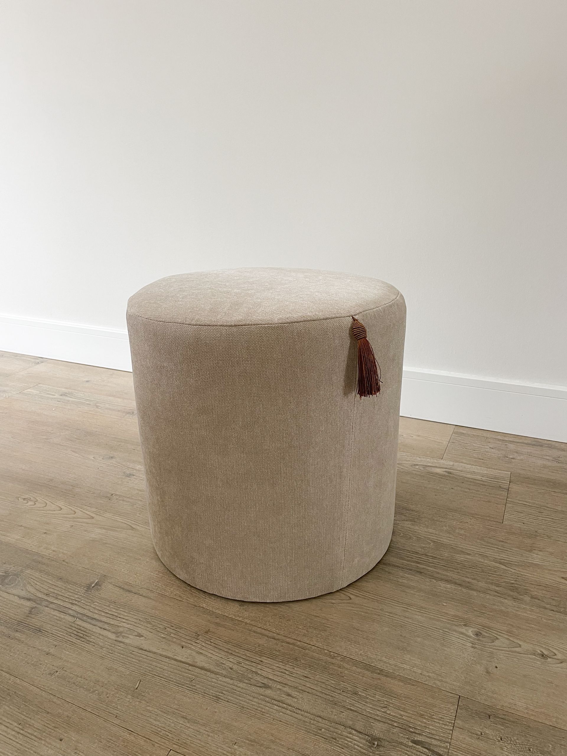 Crèmekleurige, gestructureerde, cilindrische poef op een lichte houten vloer tegen een witte muur, met een klein bruin stoffen labeltje.