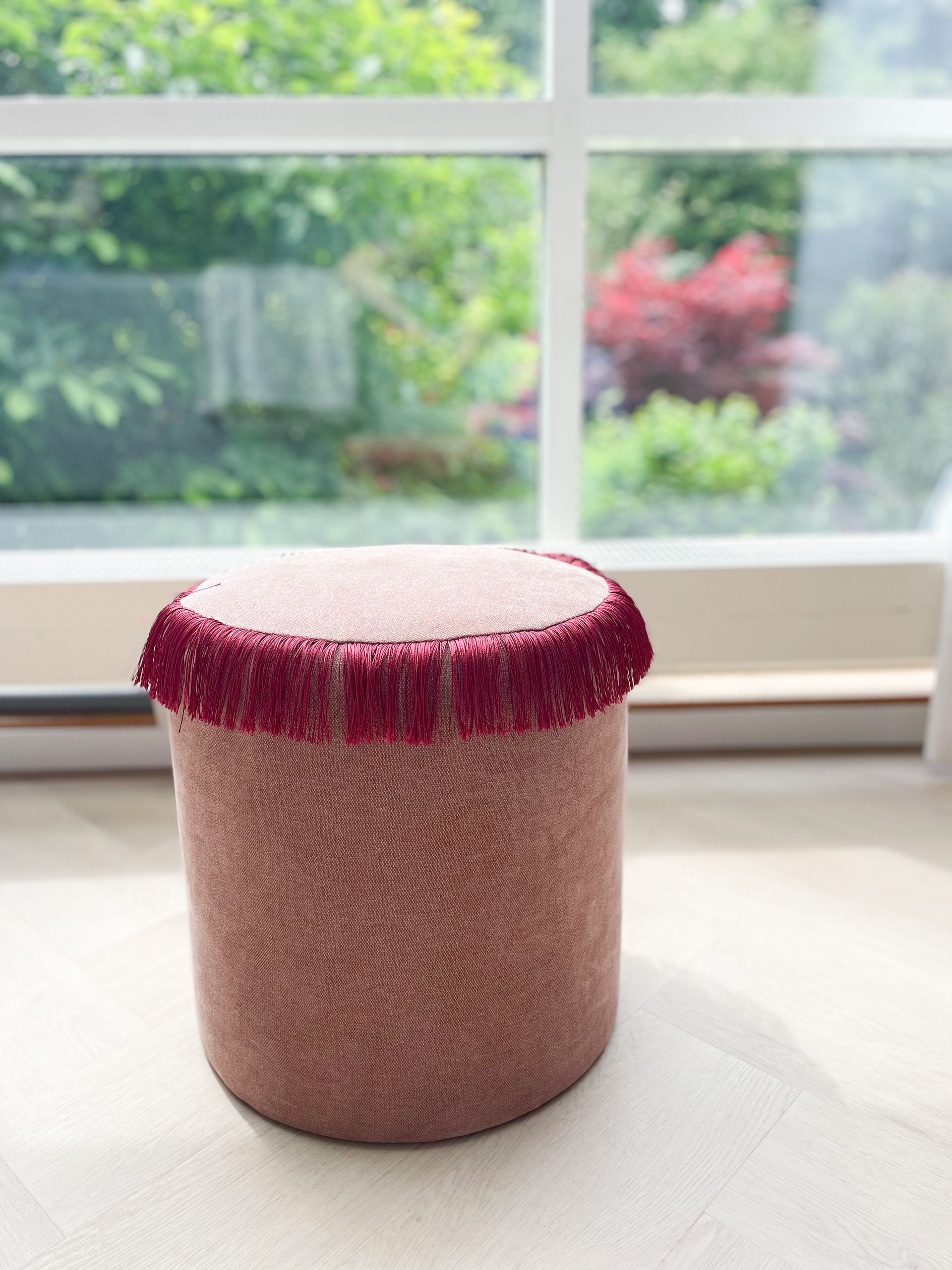 Roze, gestructureerde poef met bordeauxrode franjes voor een licht raam.