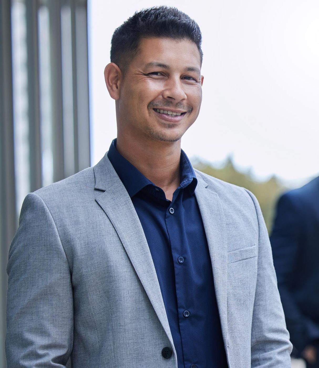 A smiling person wearing a dark blue button-down shirt and a grey textured blazer, standing outdoors.