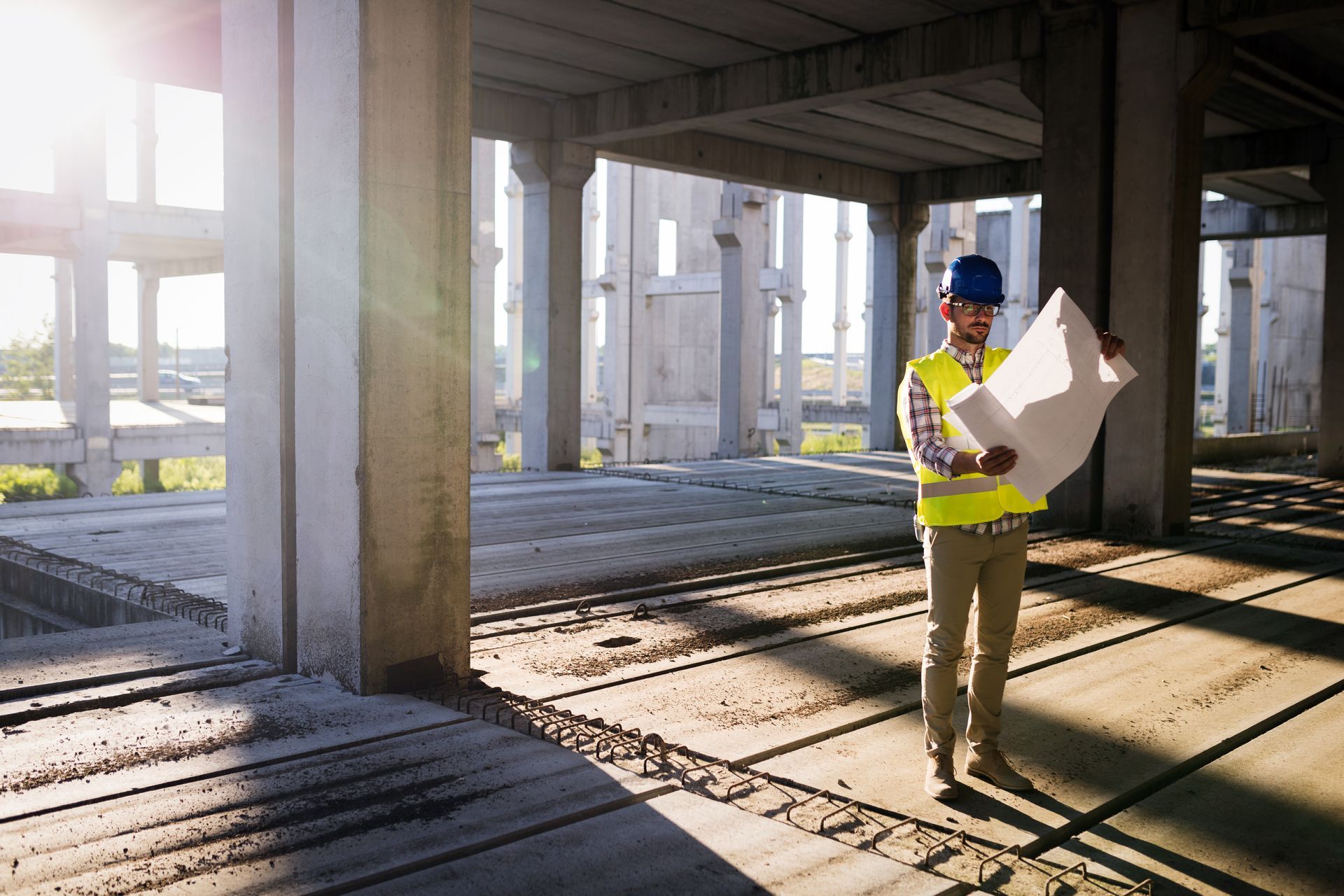 A construction worker in a yellow safety vest and blue hard hat stands in an unfinished concrete building reviewing plans.