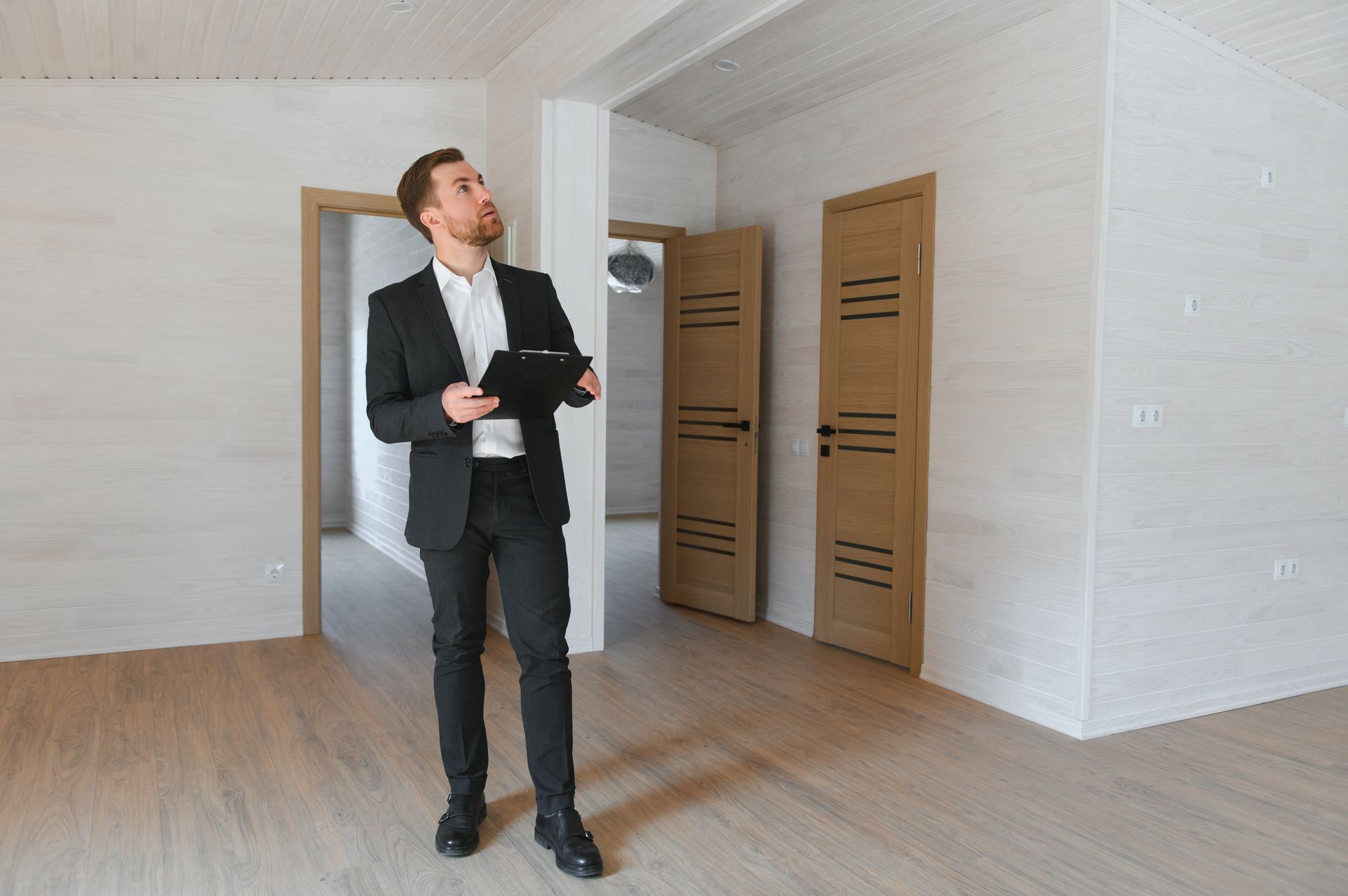 A person in a business suit holding a clipboard, standing in an empty, light-colored room and looking toward the ceiling.