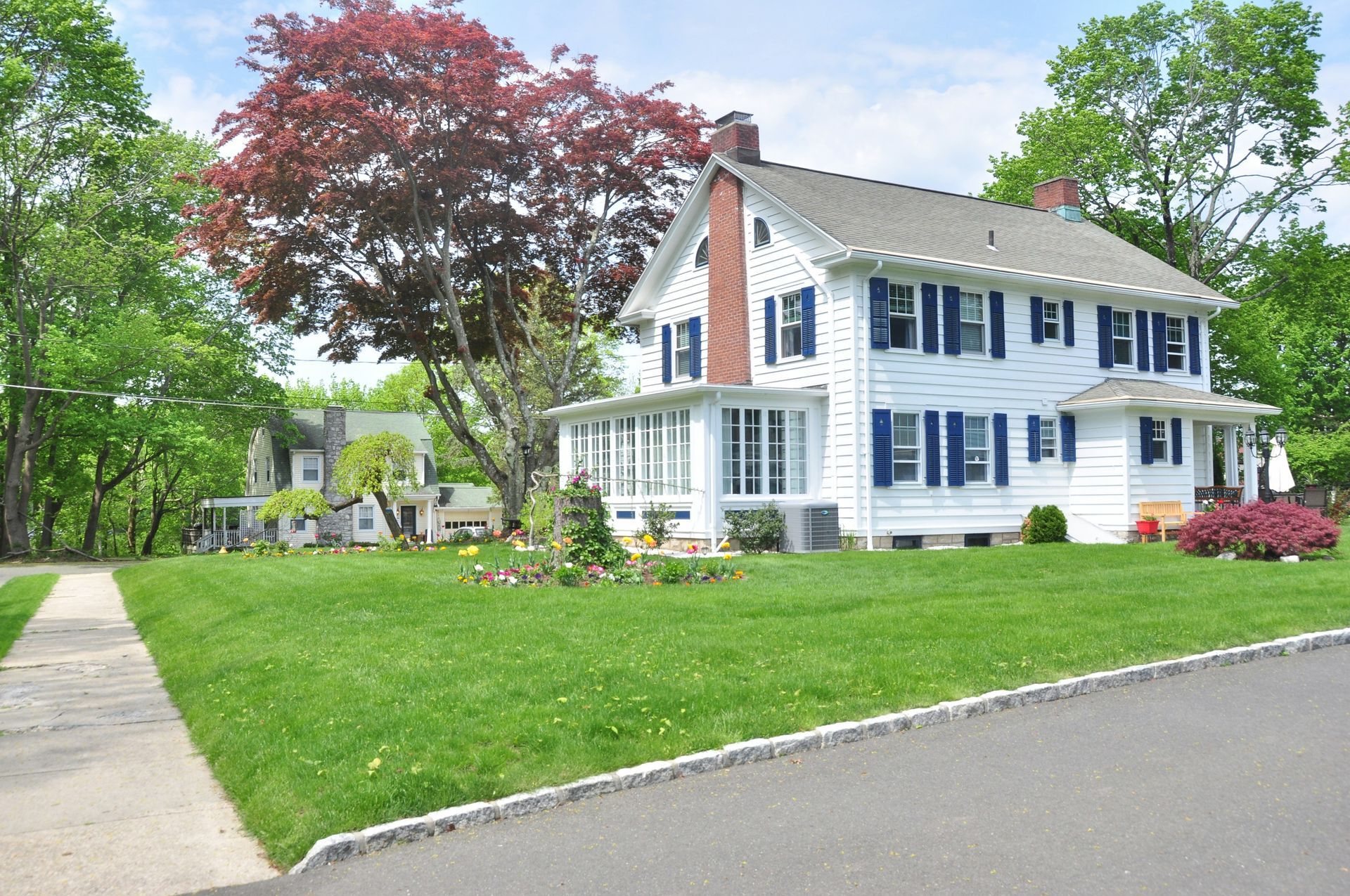 A white two-story house with blue shutters, a brick chimney, and a front porch, set on a grassy lawn with trees.