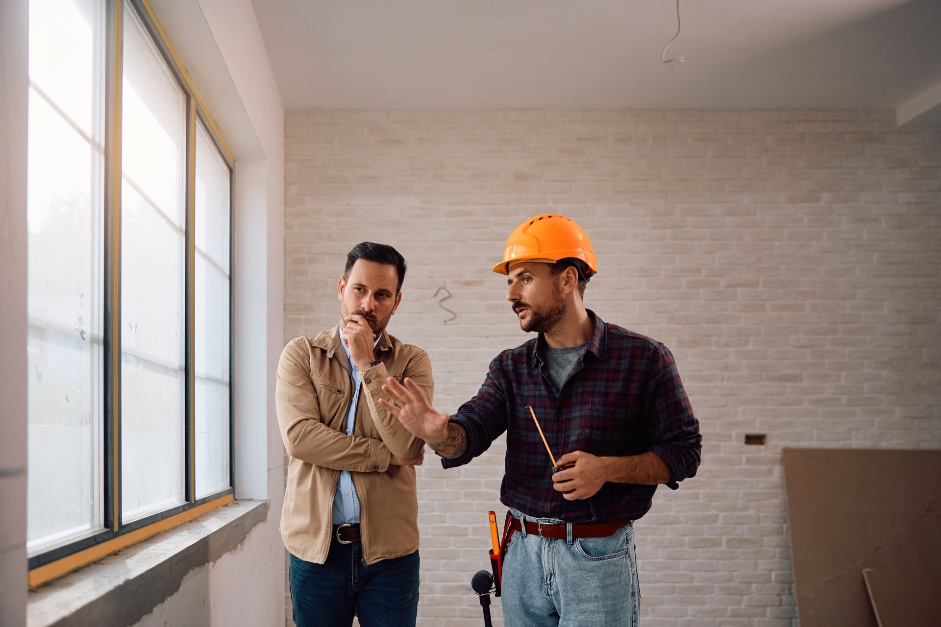 A contractor in a hard hat gestures to a window, explaining construction plans to a person in an unfinished room.