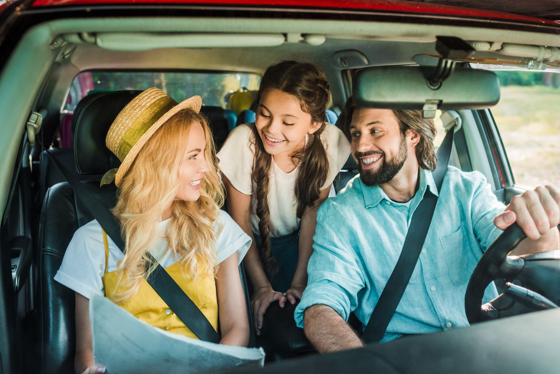 A family of three smiling inside a car during a road trip, with the driver at the wheel and others looking at a map.