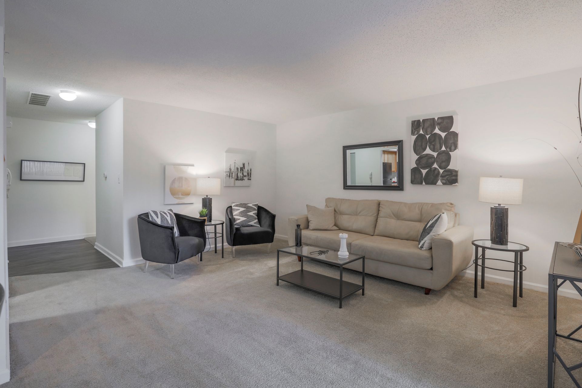 Spacious neutral living room with beige sofa, two gray chairs, coffee table, lamp, and wall art on white walls.