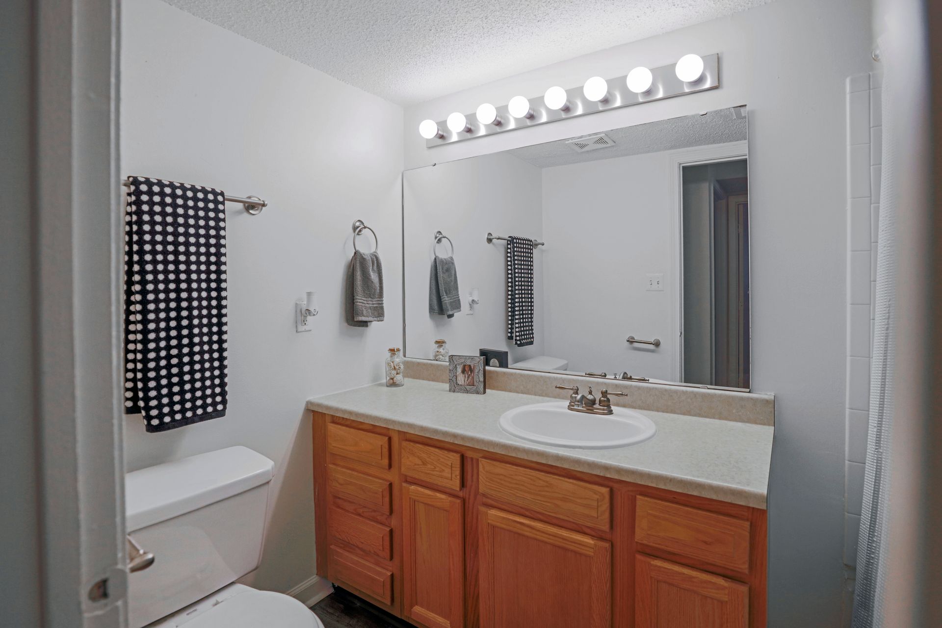 Bright bathroom with wooden vanity, large mirror, lights, and a toilet beside a towel rack.