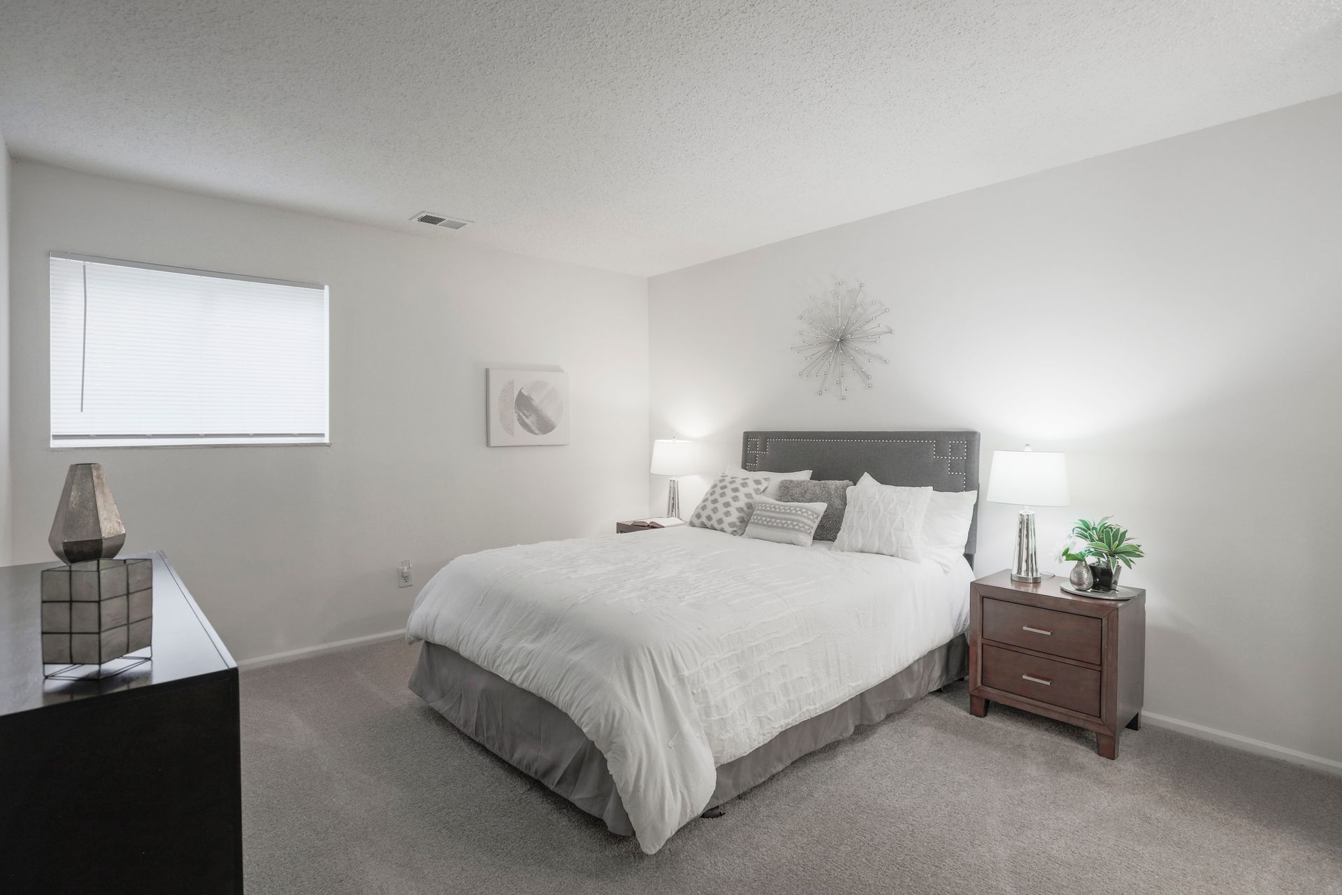 Bright bedroom with a gray bed, white bedding, nightstands, and a window with blinds.