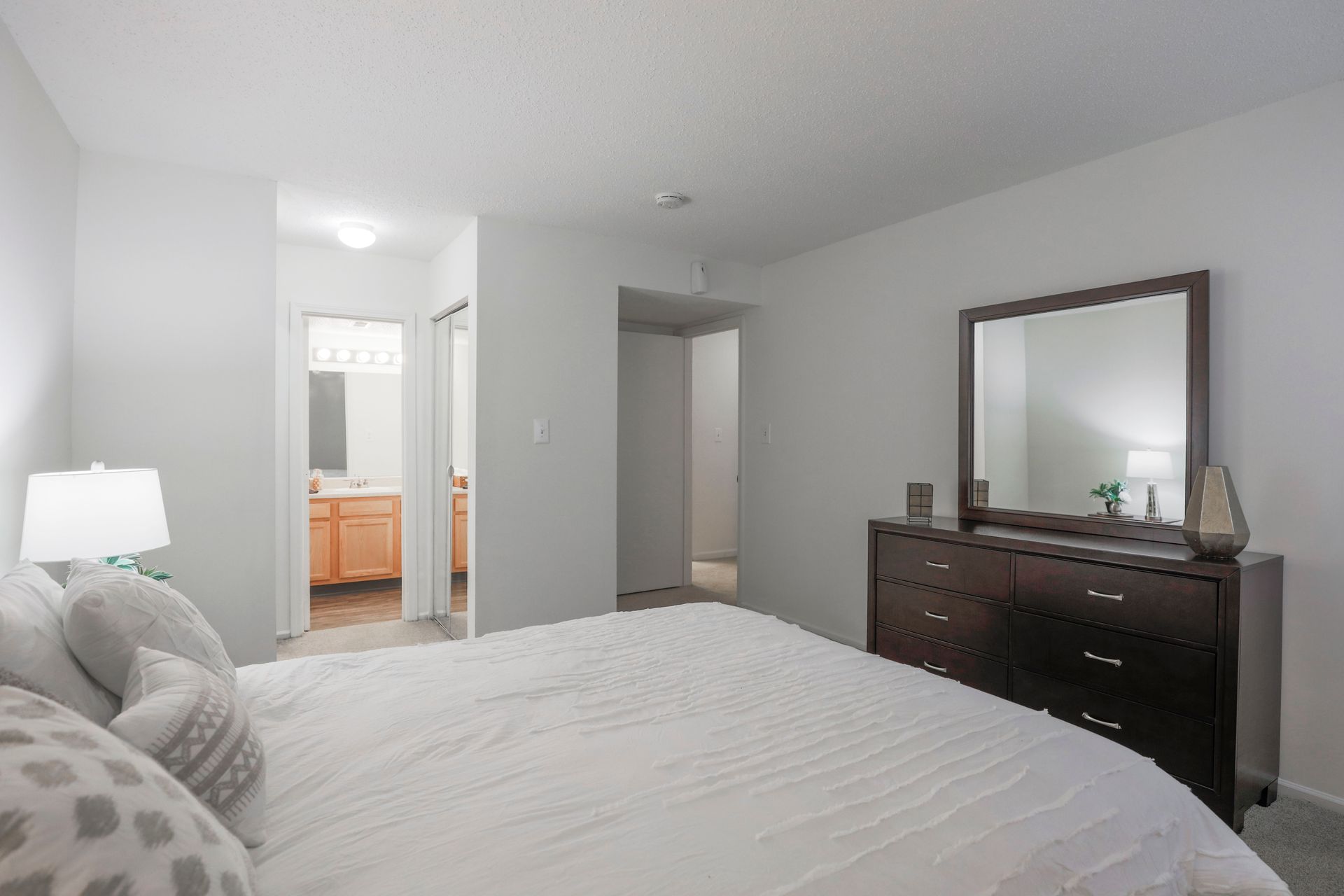 Bright bedroom with white bedding, dark dresser and mirror, and an open doorway to a bathroom.