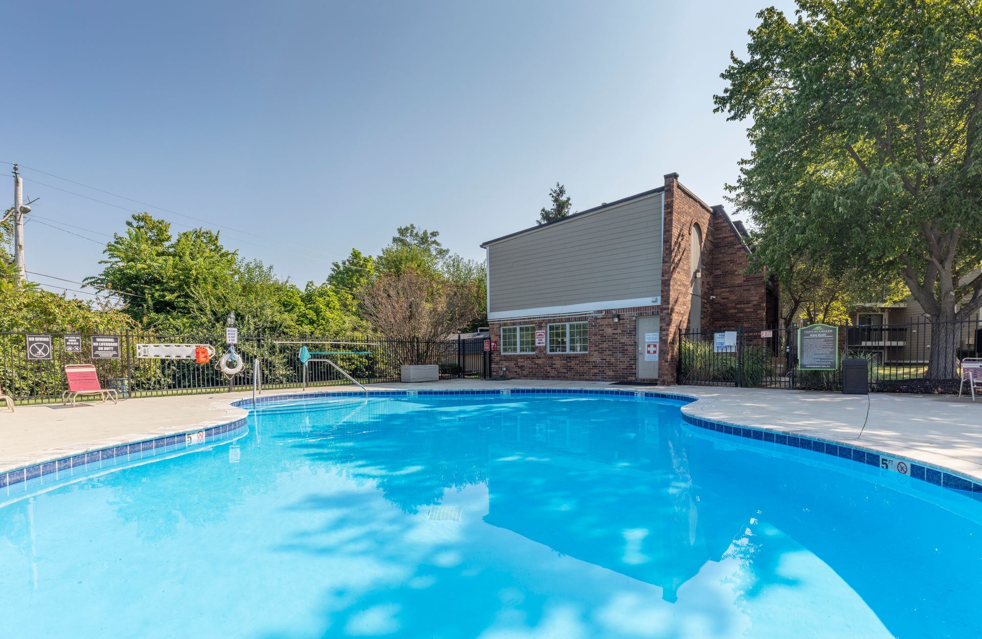 Outdoor swimming pool with clear blue water beside a brick building and trees on a sunny day