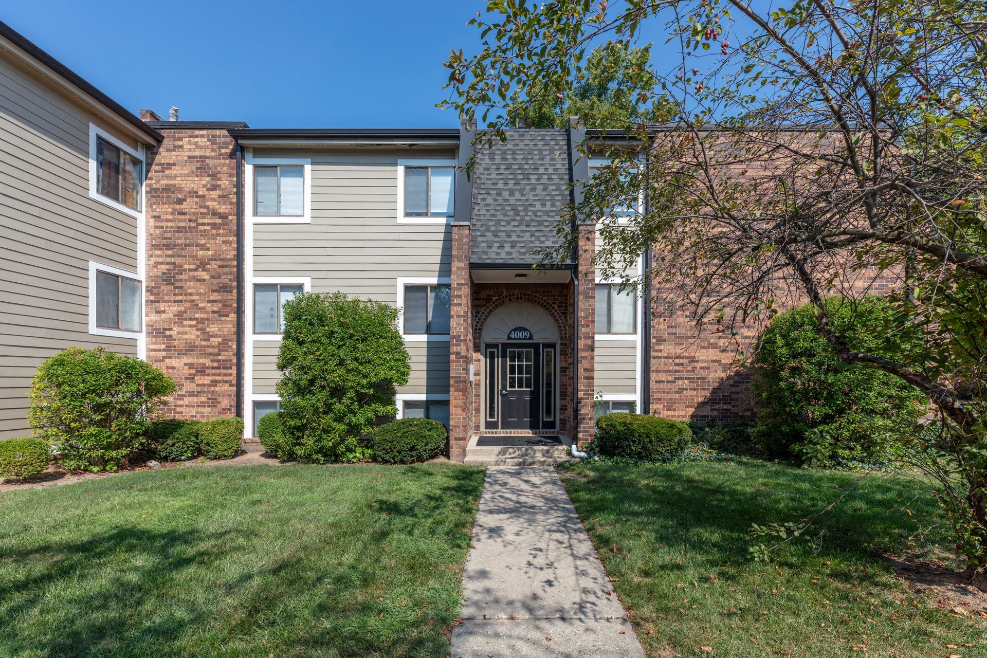 Apartment building entrance with brick facade, central walkway, and grassy front lawn