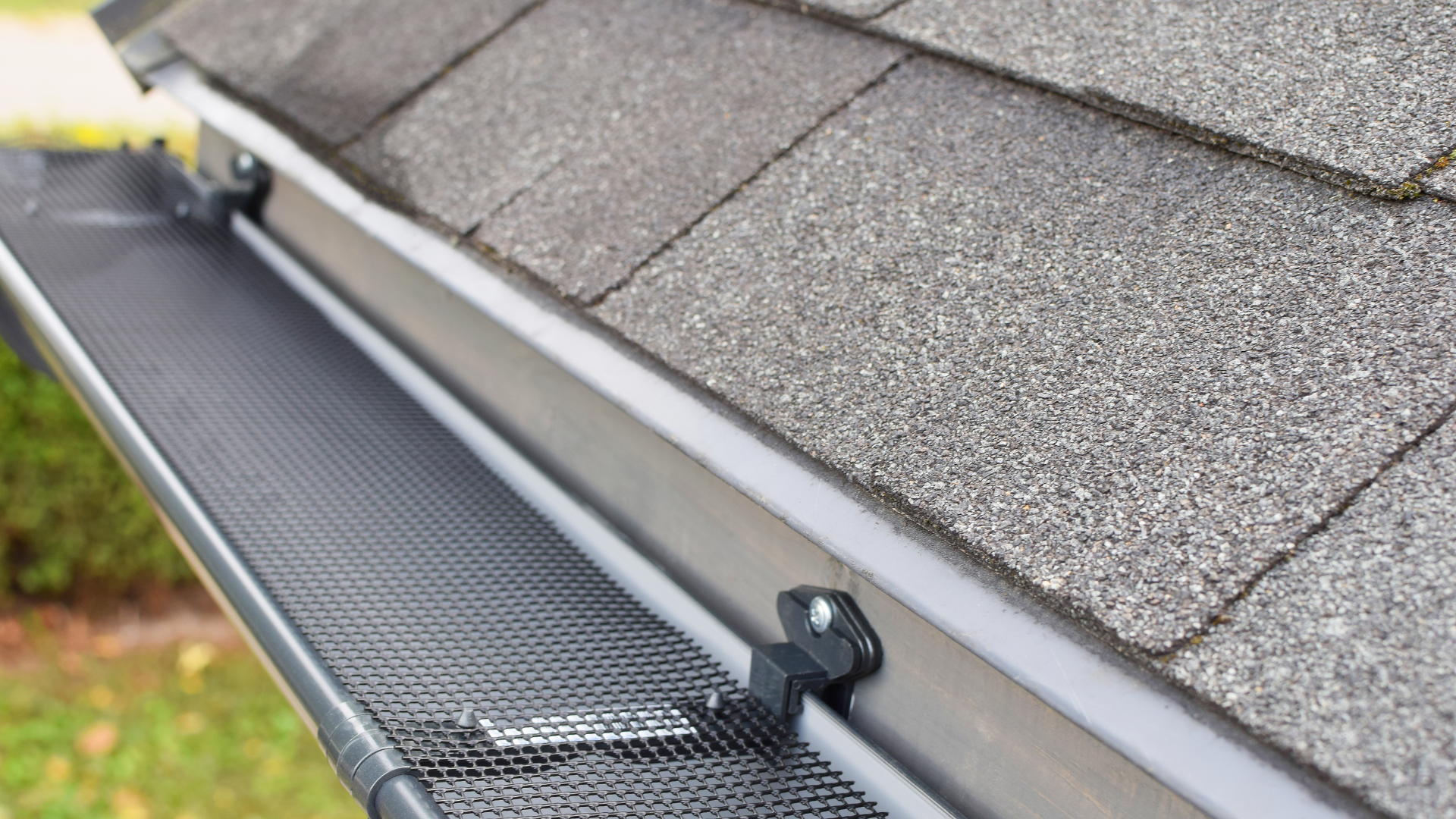 Gutter guard installed along a roof edge beside gray asphalt shingles and a downspout bracket.