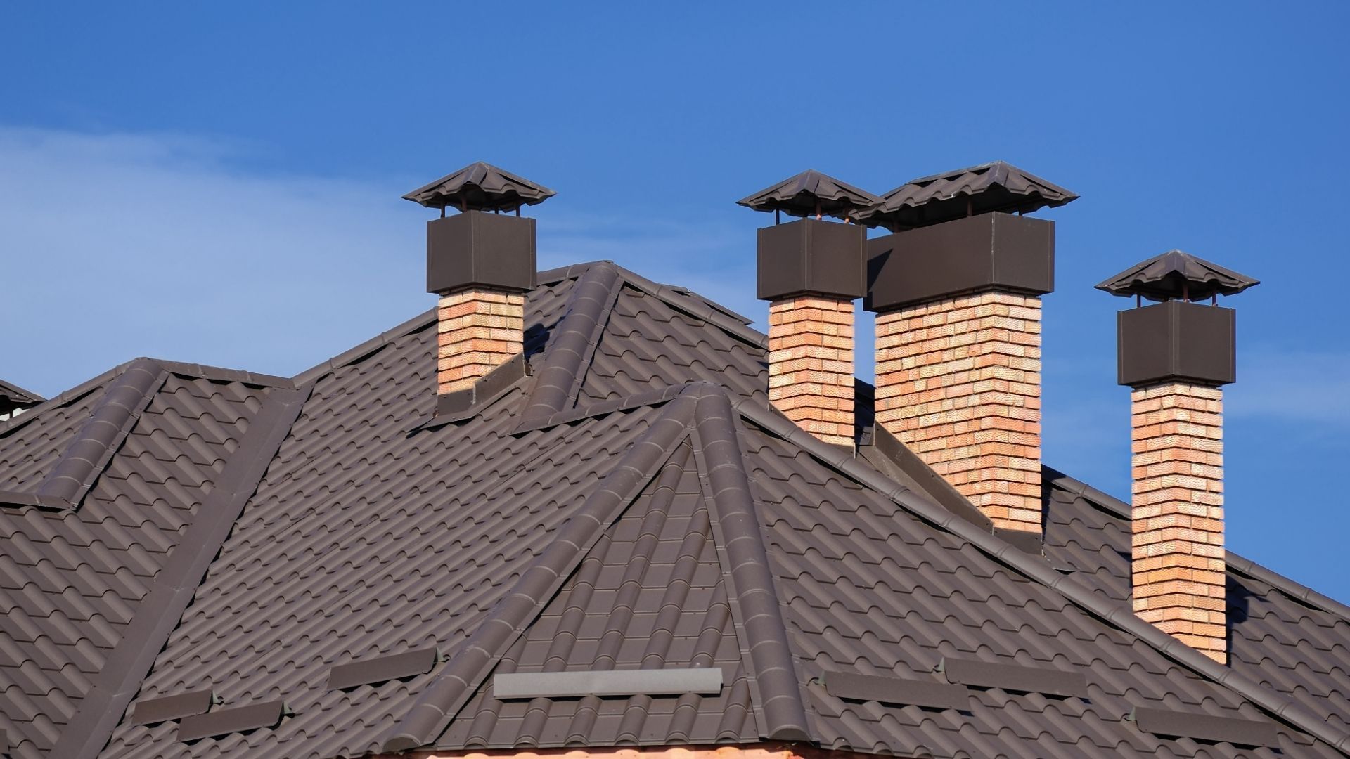 Dark shingled roof with several brick chimneys against a clear blue sky