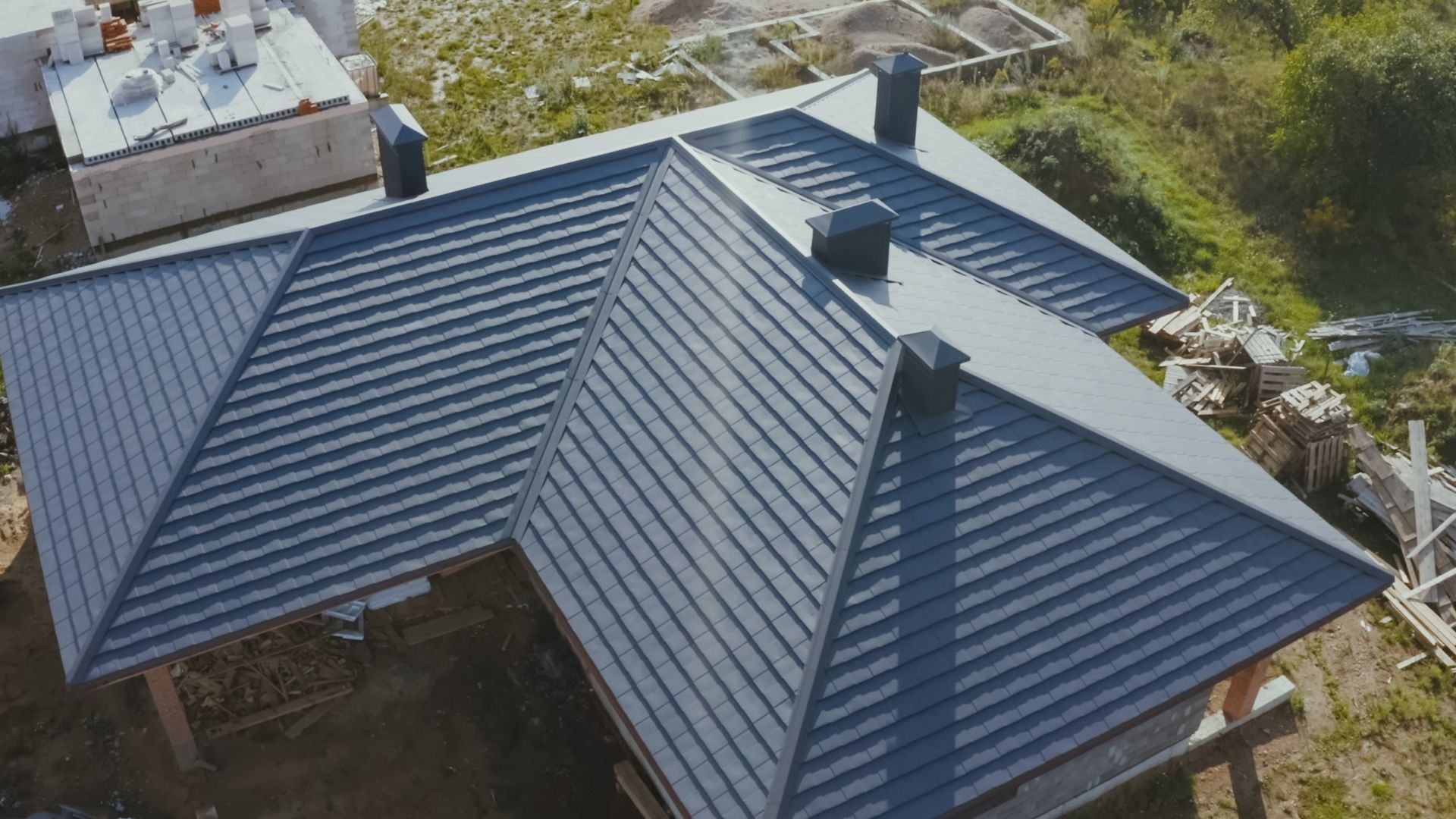 Aerial view of a house with a dark blue metal roof and skylight, surrounded by greenery.
