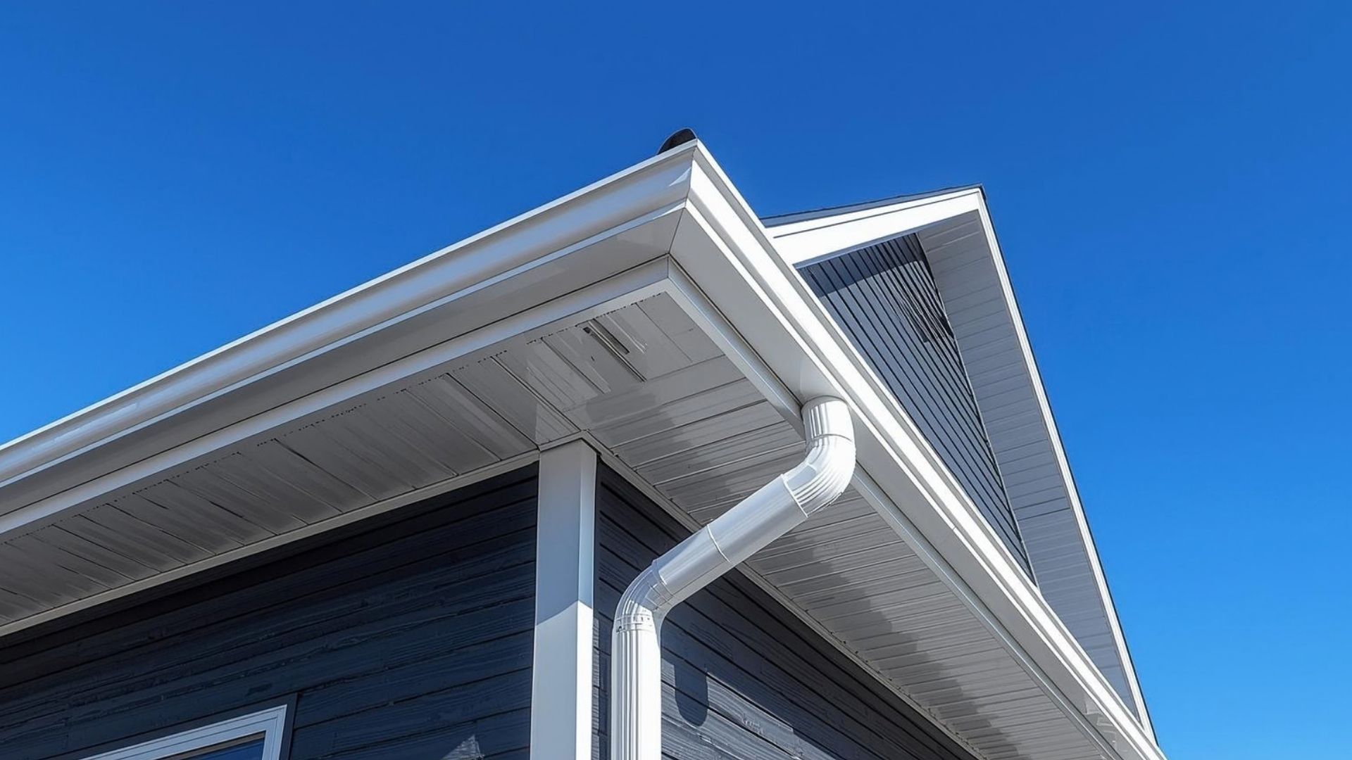White house roof eaves with a gutter and downspout against a clear blue sky