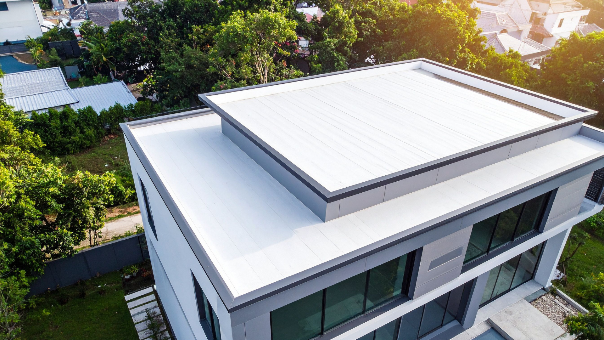 Modern white-roofed house viewed from above, surrounded by green trees and sunlight