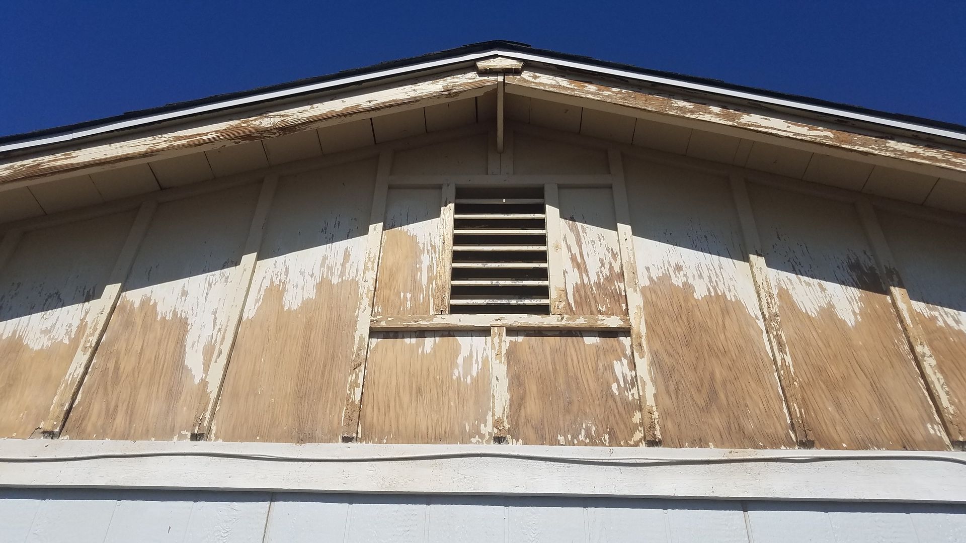 Weathered house gable with peeling tan siding and a small vent under the roof peak.