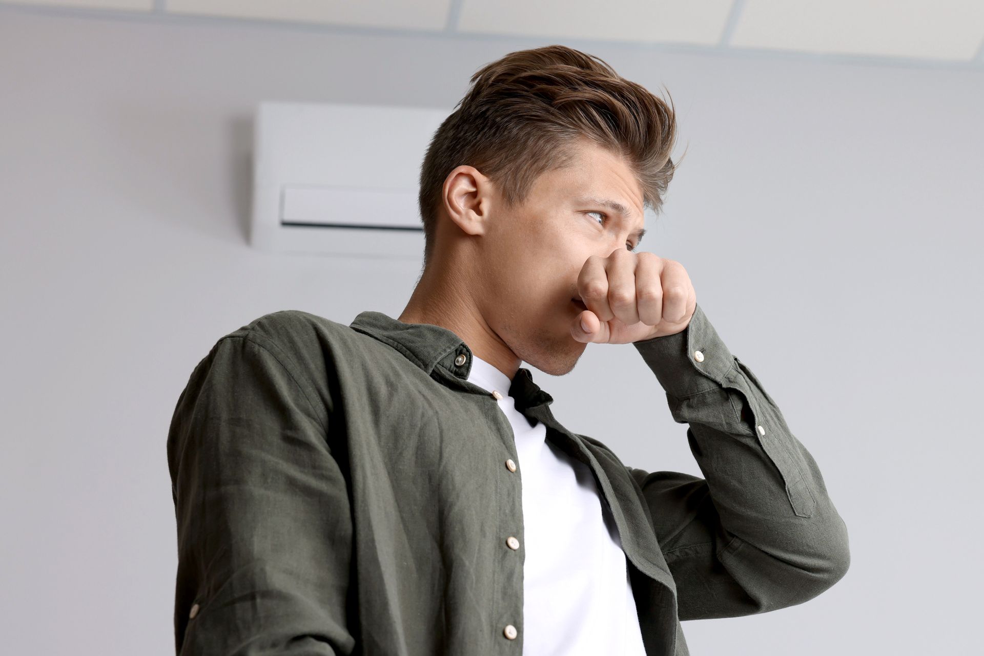 Man in green shirt coughing into his fist indoors, with a wall-mounted air conditioner behind him