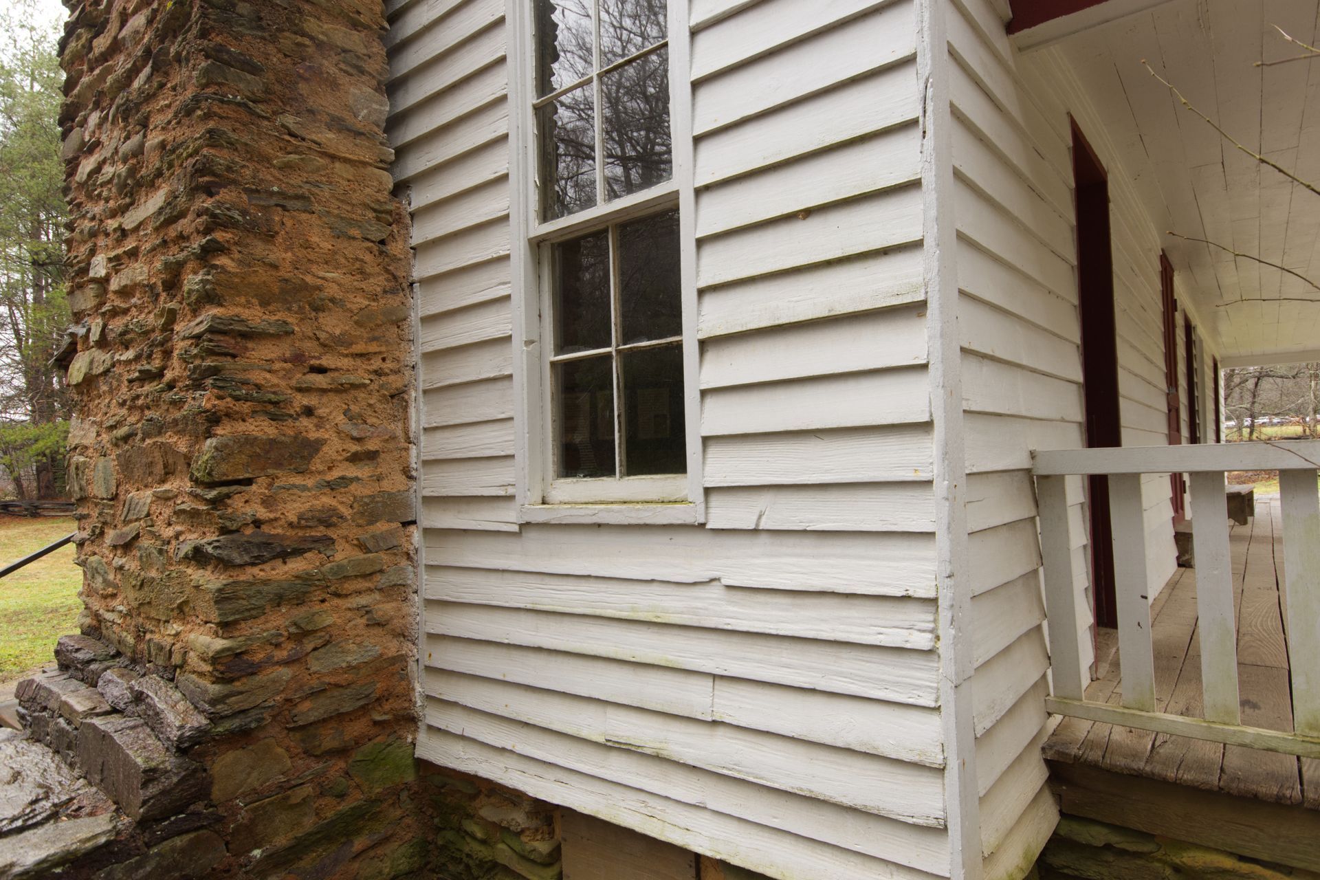 Side of a white house with a stone chimney and porch railing in a grassy yard
