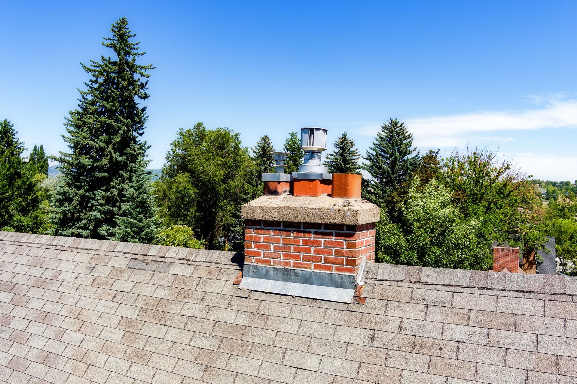 Brick chimney on shingled roof with metal vents, against green trees and blue sky