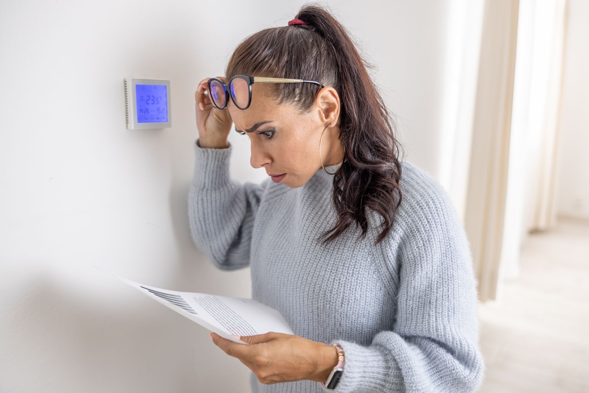 Woman reading a document beside a wall thermostat, holding glasses in a bright hallway