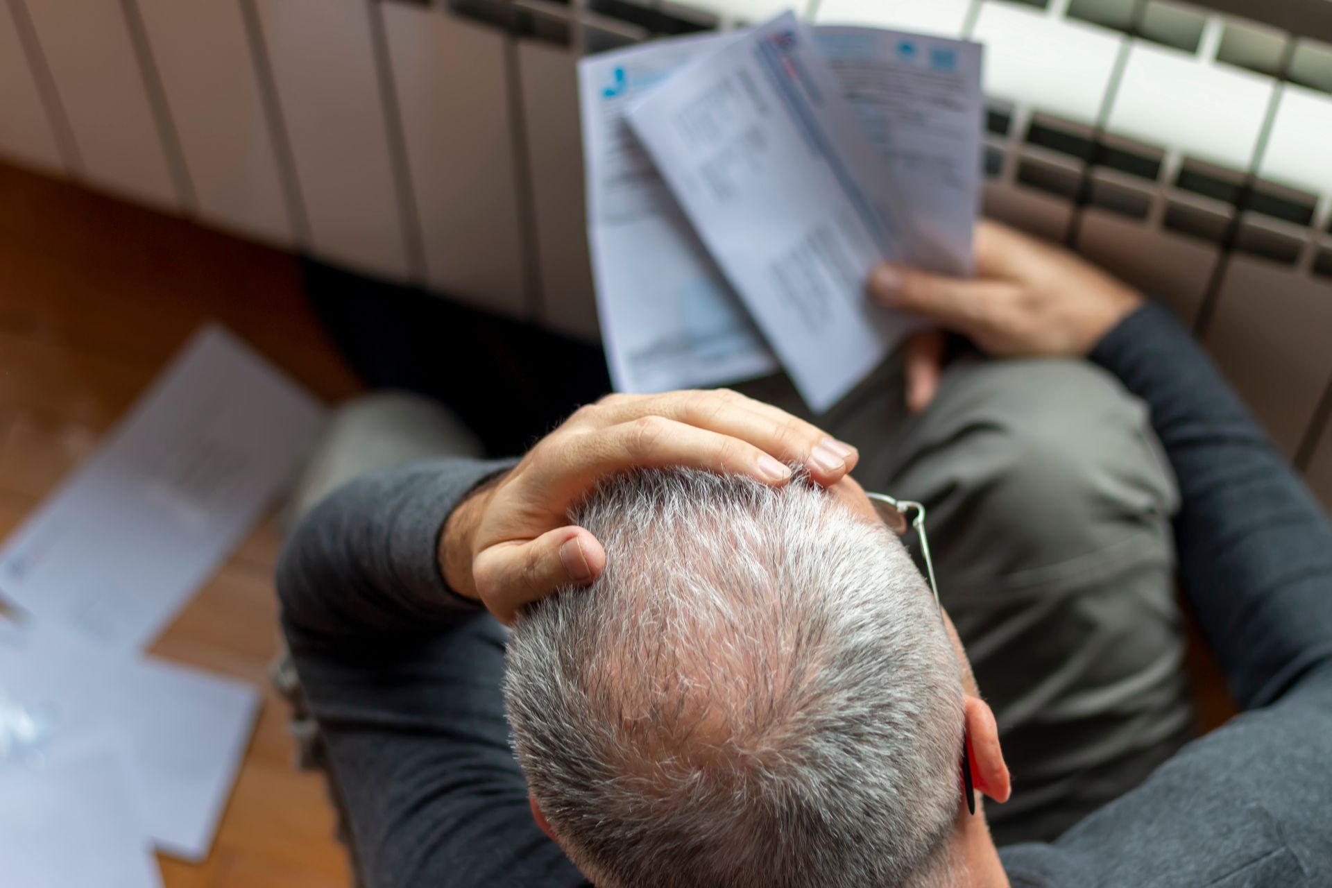 Person sits on a couch holding their head and reading papers on the floor.