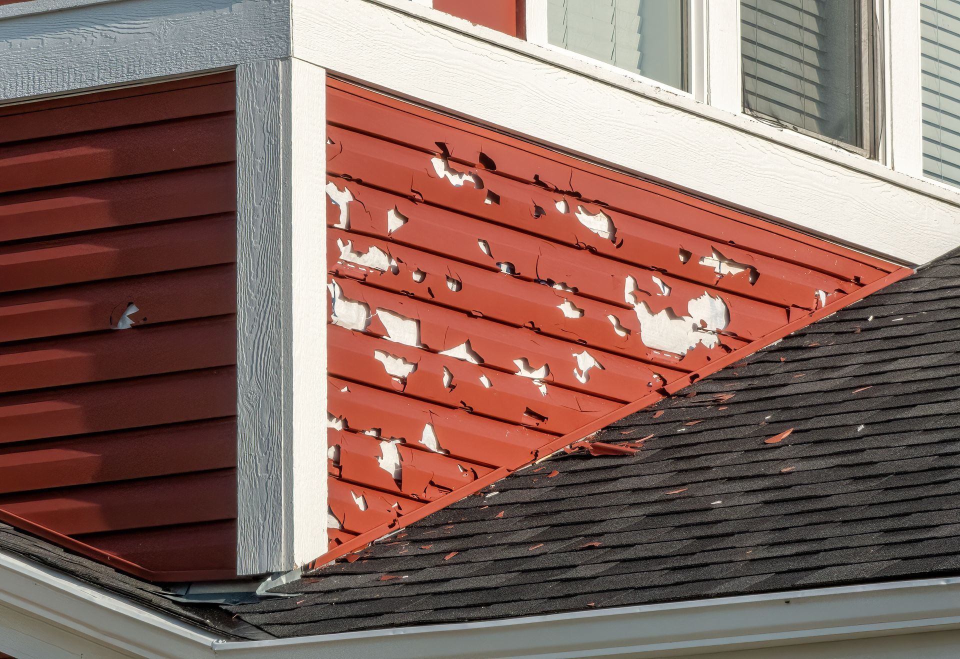 Peeling red siding on a house exterior above a shingled roof and white trim