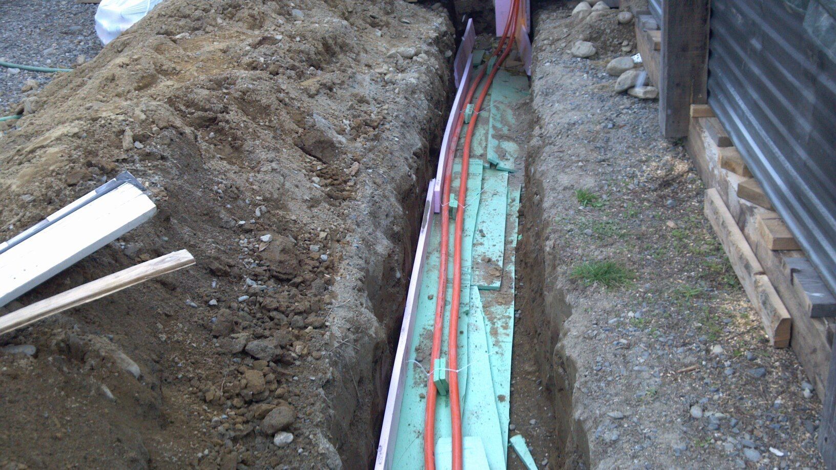 Trench beside a building with exposed red cables and turquoise conduit running along the ground
