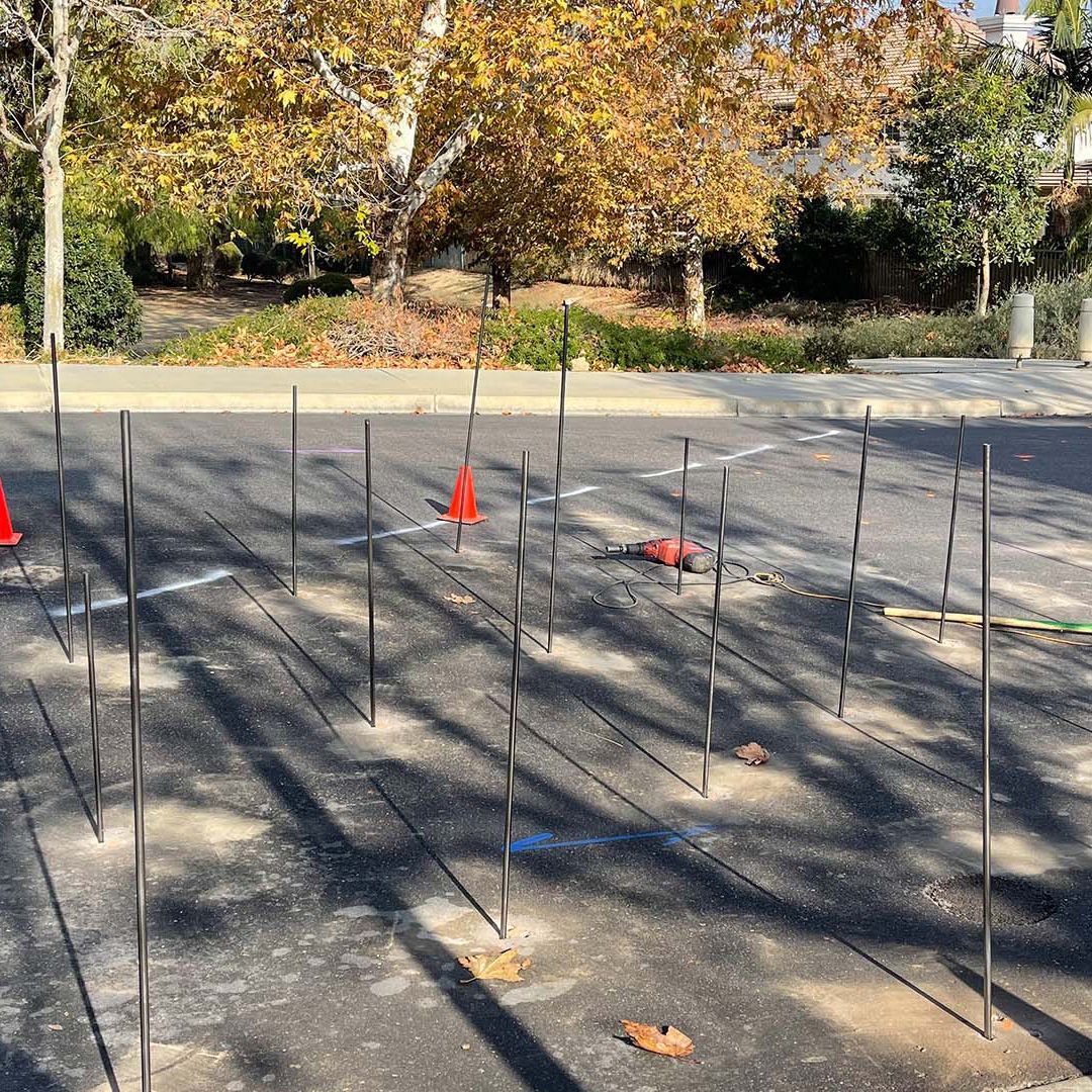 Driveway with many thin stakes or wires laid out in rows, marked by orange cones under autumn trees.