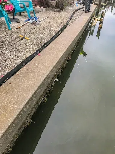 Concrete walkway beside a murky canal, with turquoise chairs and a black hose along the edge.