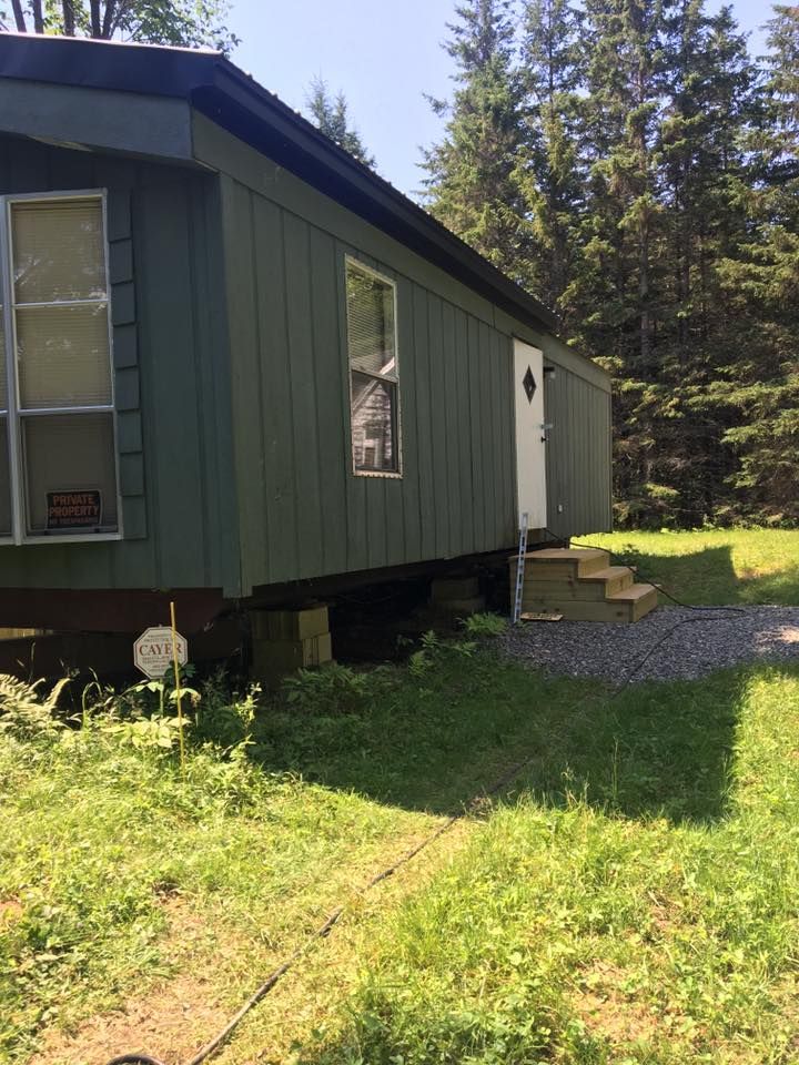 Green cabin with a white door on a grassy lot, with trees in the background.