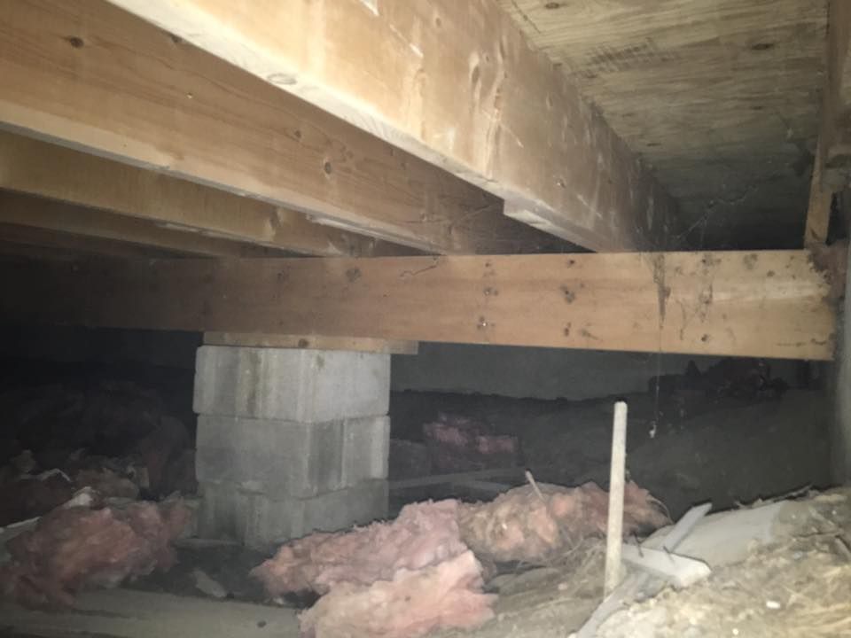 Crawlspace under a wooden floor with support beams, cinder block pier, and pink insulation on the ground