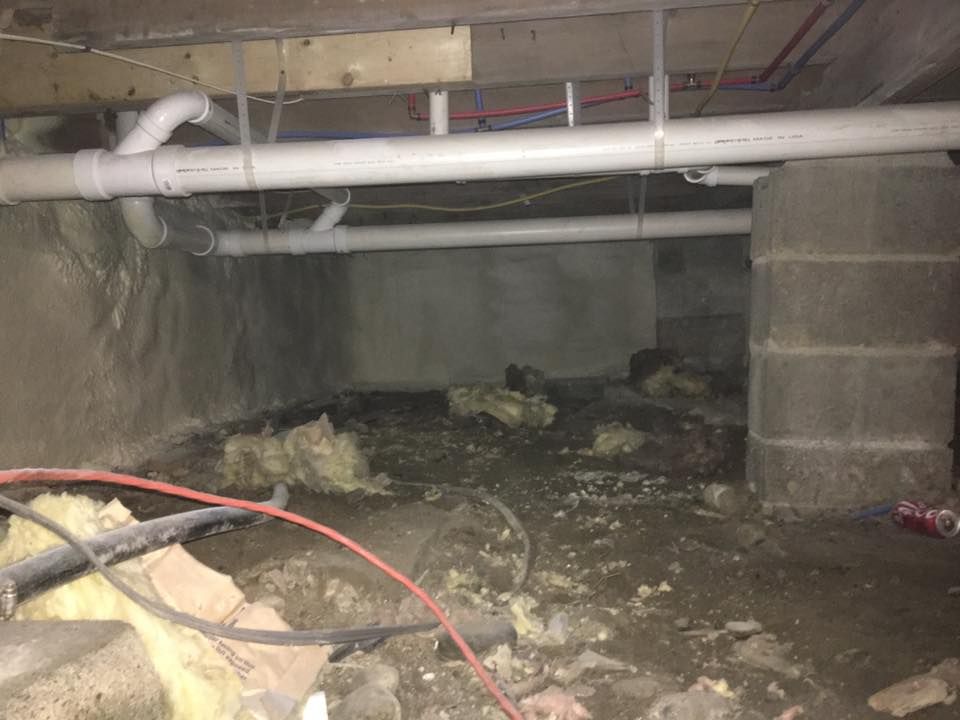 Crawlspace under a house with exposed pipes, concrete supports, and debris on the ground.