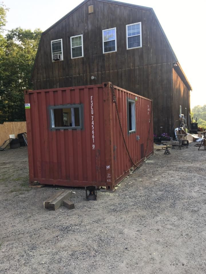 Red shipping container cabin beside a tall wooden building on a gravel lot