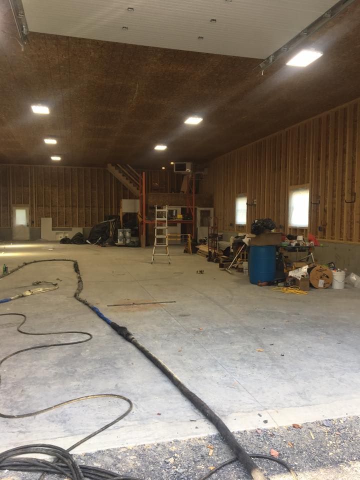 Empty workshop with concrete floor, wood-paneled walls, overhead lights, and ladders and tools at the back.