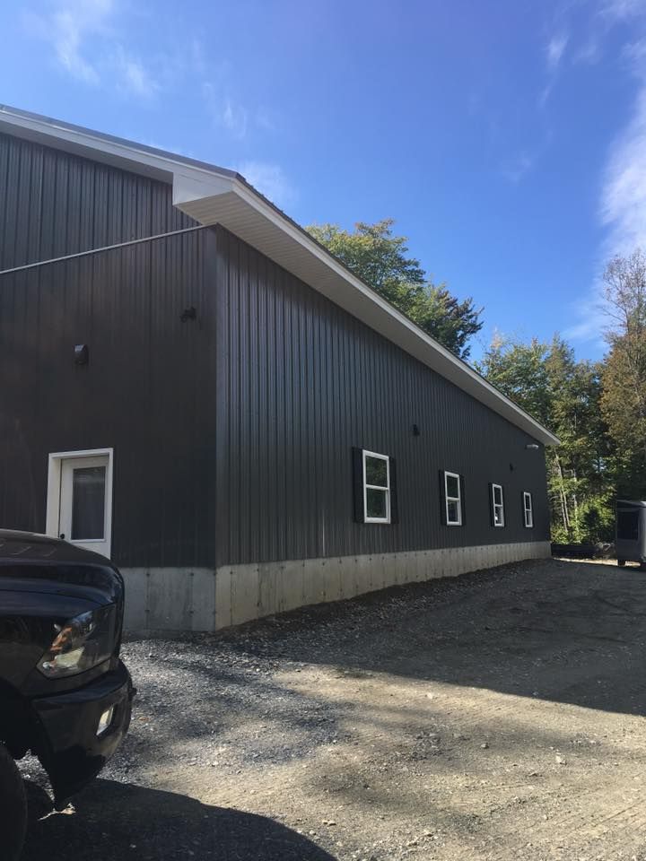 Gray metal building with small windows, gravel lot, and a black car under a blue sky.