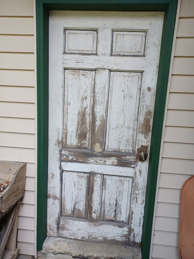 Worn exterior door with peeling white paint and visible wood damage, framed by green trim and tan vinyl siding.