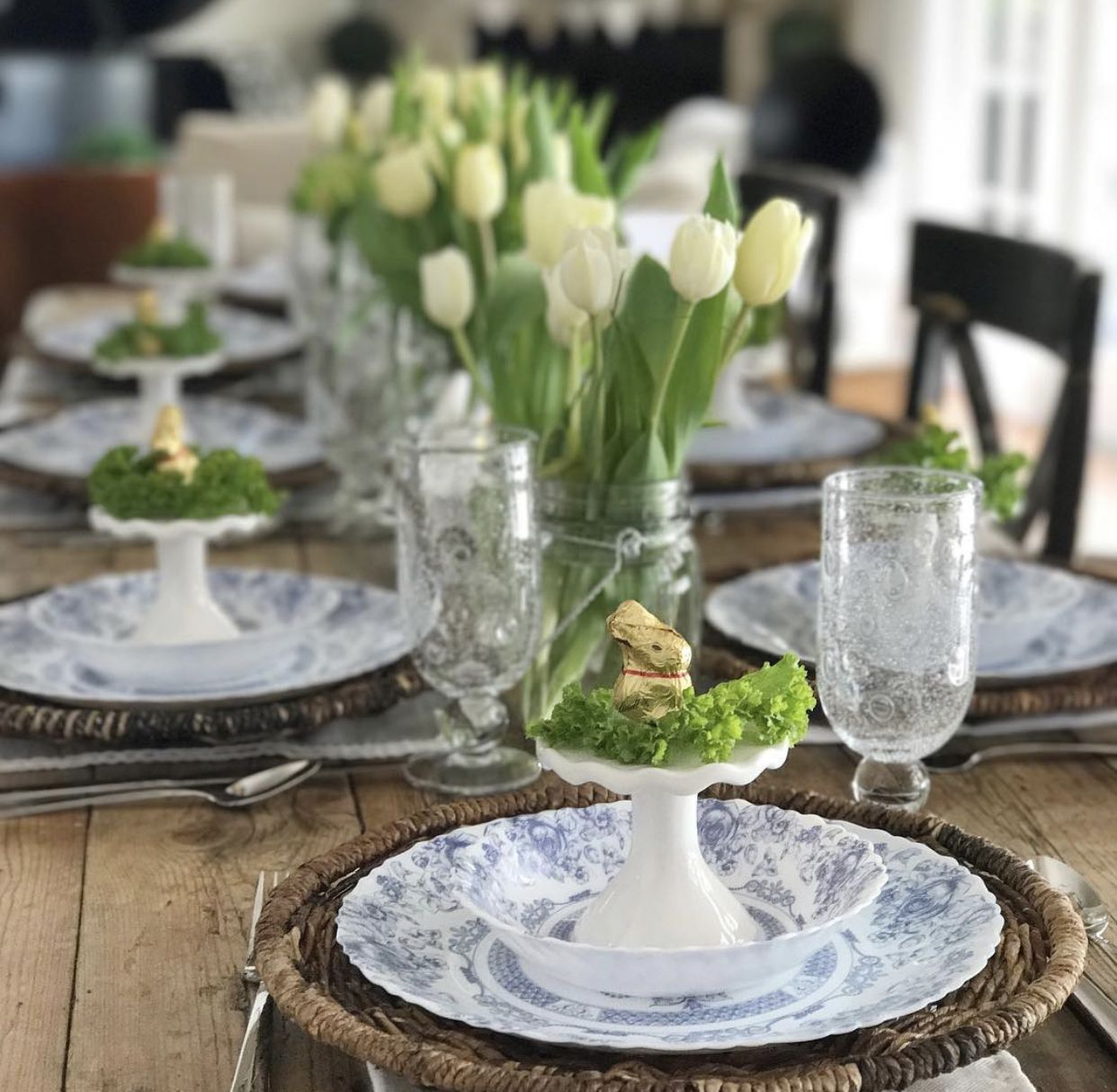 A long table is topped with Easter decor, including white tulips, blue and white china, and a chocolate Easter bunny.