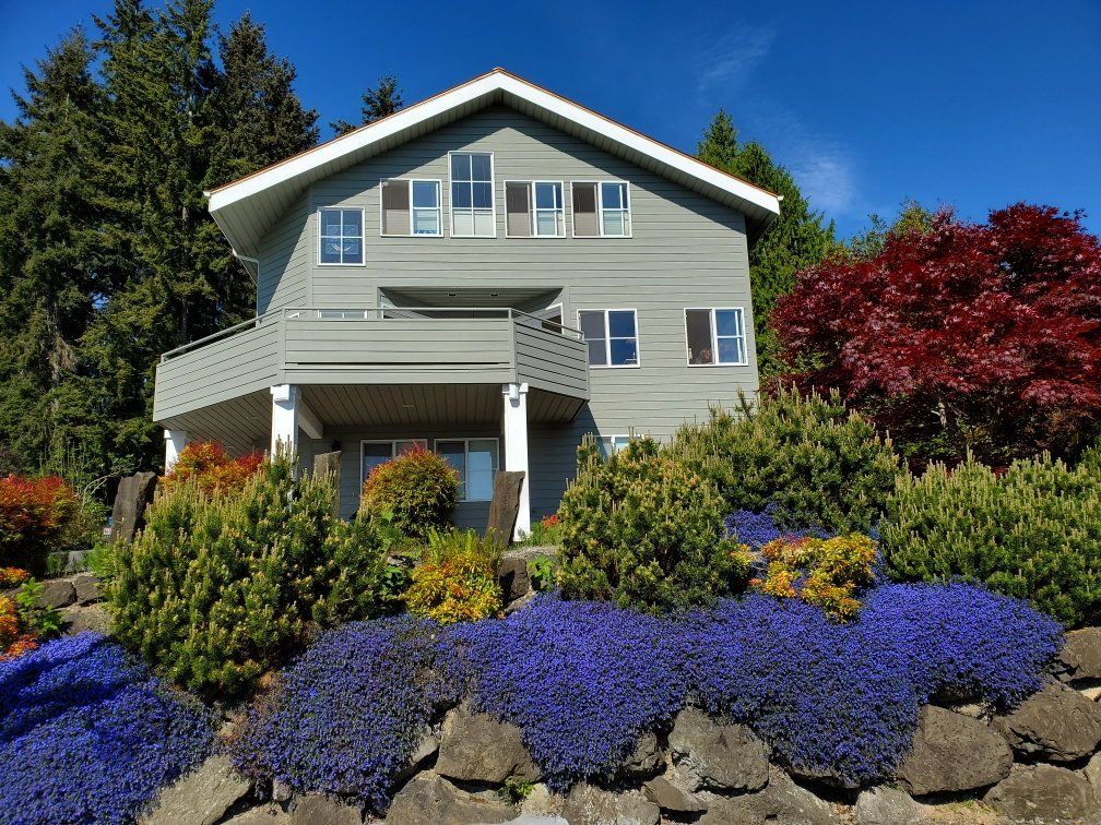 Two-story gray house with a balcony, surrounded by colorful bushes and flowers on a sunny day.
