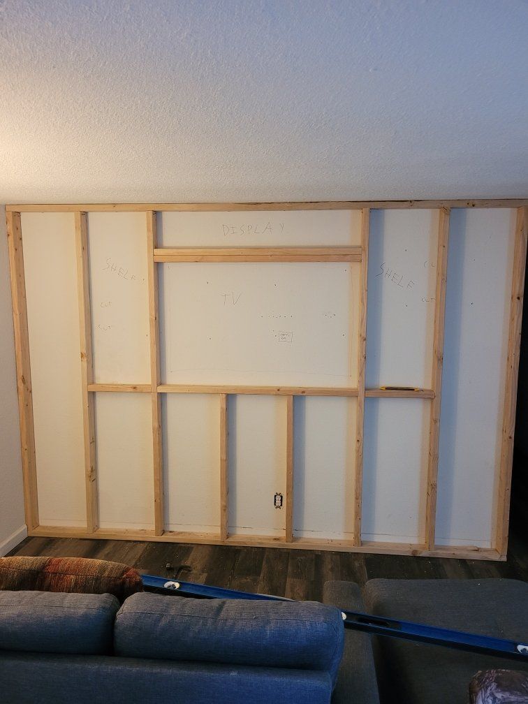 Unpainted framed wall with built-in sections for a TV, display shelf, and shelving, under construction in a living room.