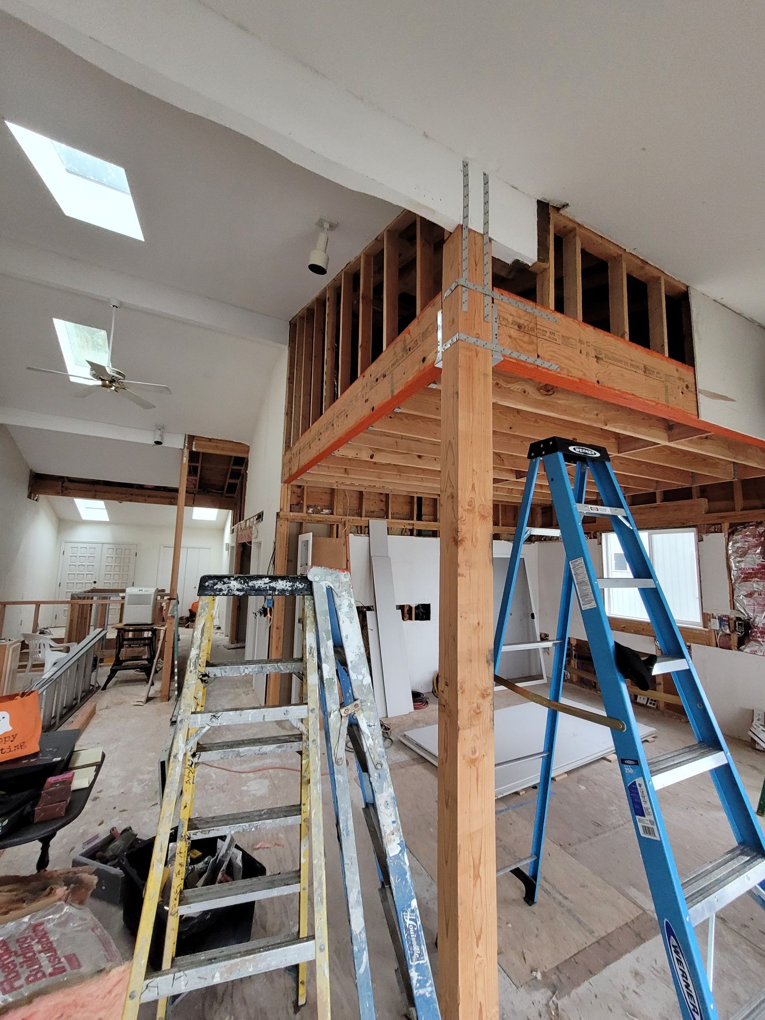 Room under renovation with exposed framing, stacked drywall sheets, and ladders set up beneath high ceilings and skylights.