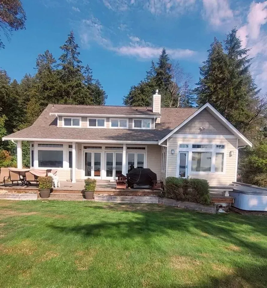 A beige house with a porch, green lawn, and tall evergreen trees under a blue sky.