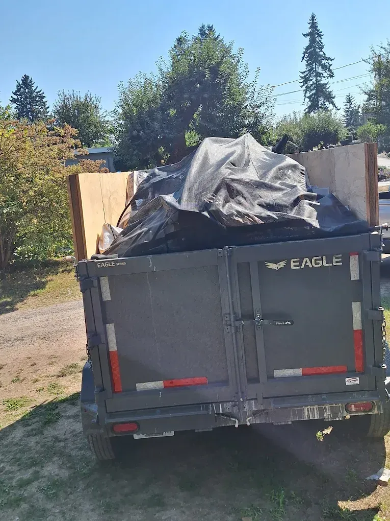 Black dump trailer loaded with debris covered in a black tarp on a sunny gravel driveway.