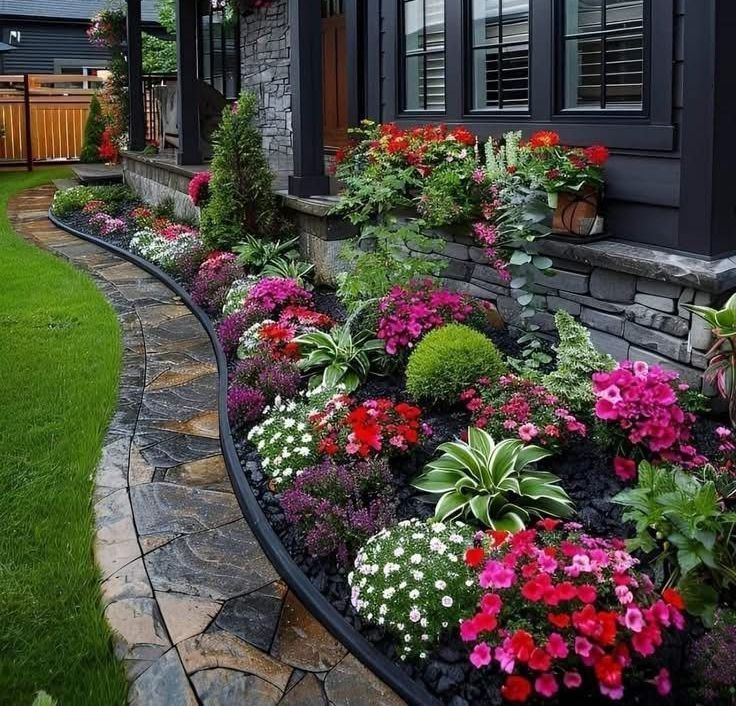 A colorful flower bed with white, pink, and red flowers lines a stone walkway leading to a dark house.