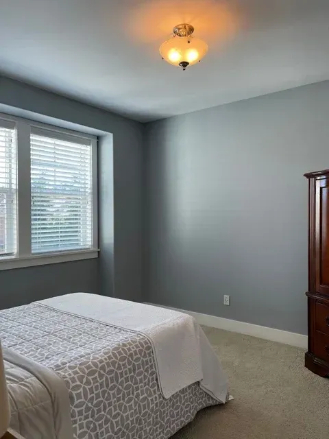 Empty bedroom with gray walls, beige carpet, white bed, window blinds, and a wooden dresser