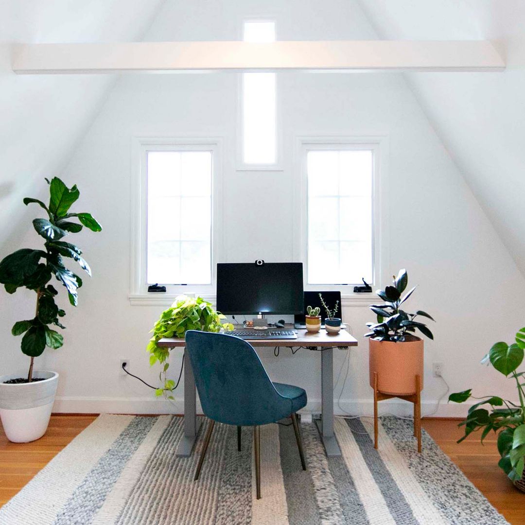 A crisp, white, a-frame room with a computer desk, symmetrical windows, and an assortment of indoor plants. 