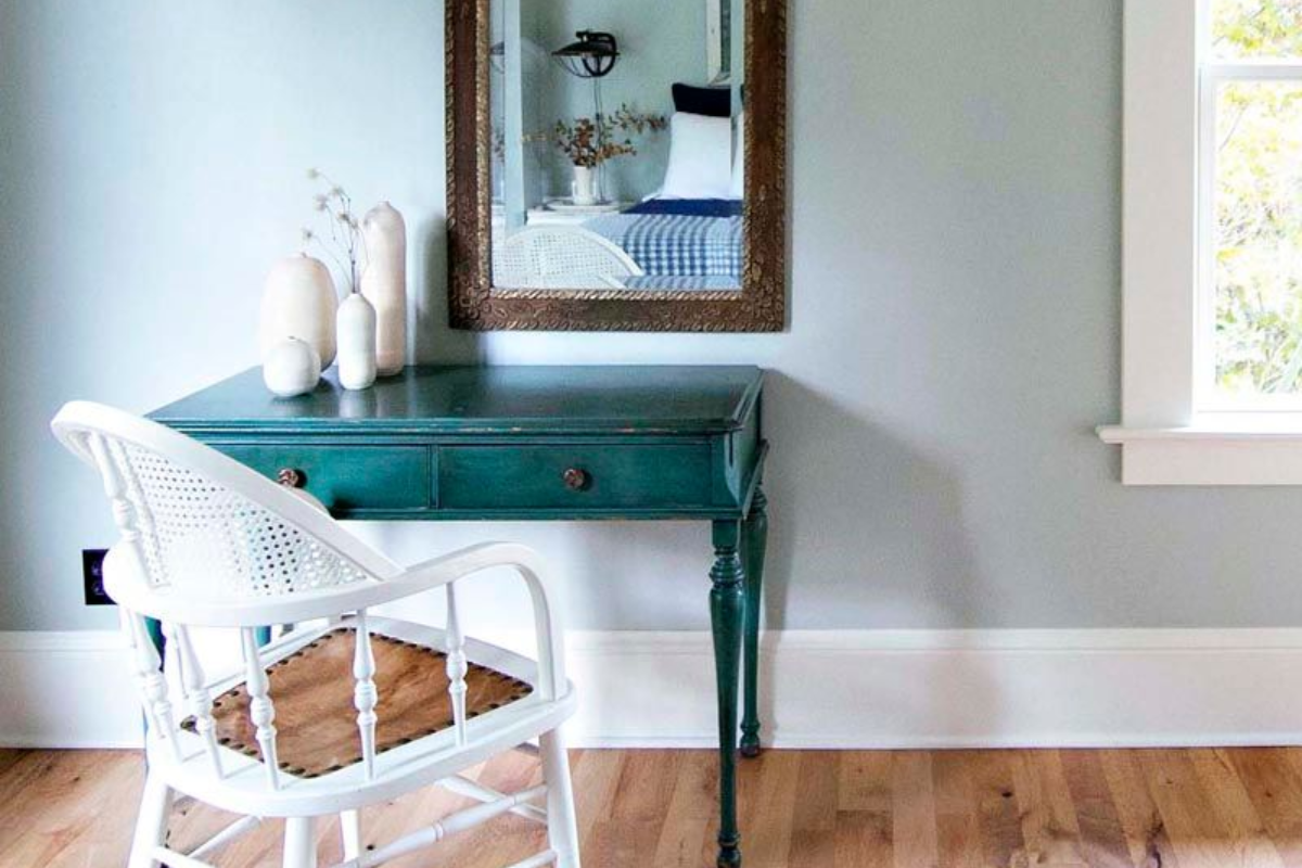 A teal desk with white chair and mirror above, against a light blue wall, white trim, and hardwood floor.