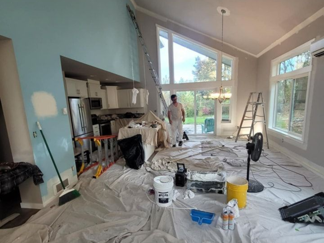 Living room with high ceilings prepped for painting with plastic-covered floors and tools.