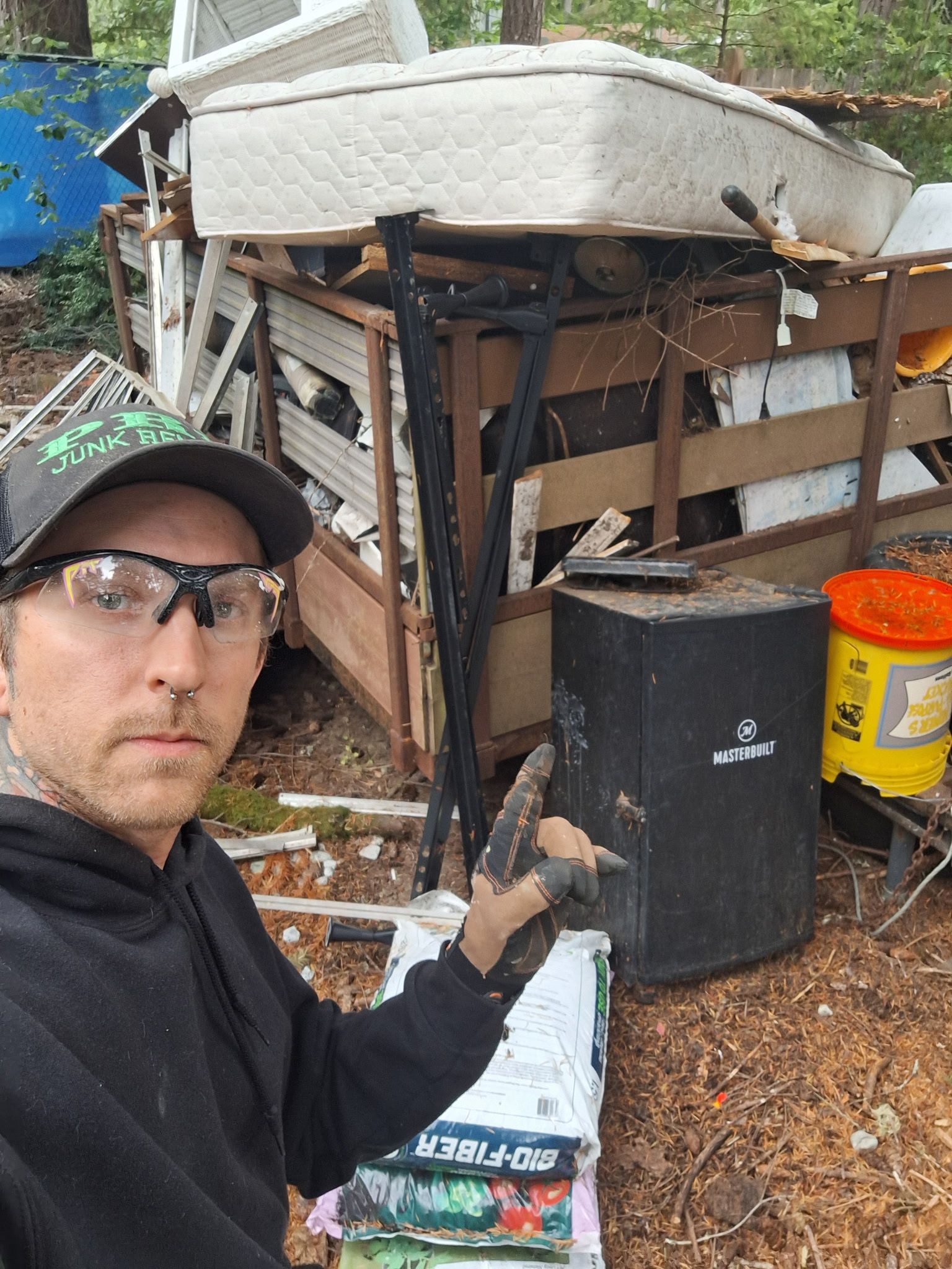 Man in safety glasses posing beside a dumpster trailer packed full with furniture, a mattress, and other household junk.
