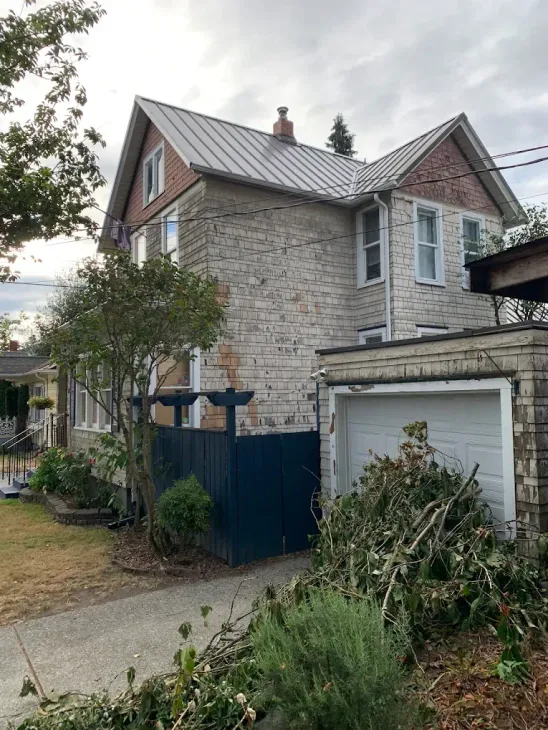 Two-story house with worn siding, a metal roof, a garage, and overgrown front garden
