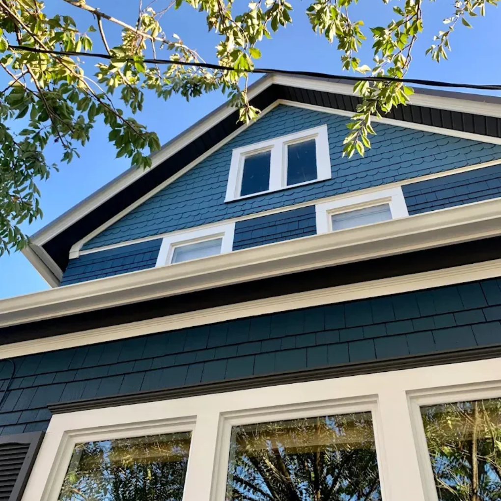 Blue house exterior with white-trimmed windows and leafy branches against a clear sky